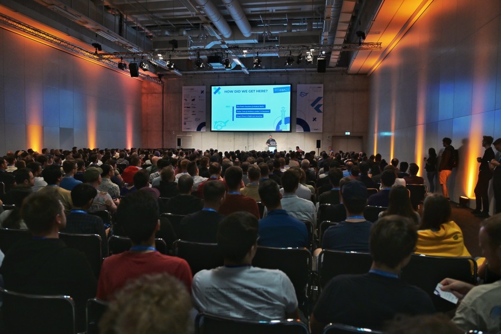 The picture shows a full exhibition hall with many people standing in front of a large screen.